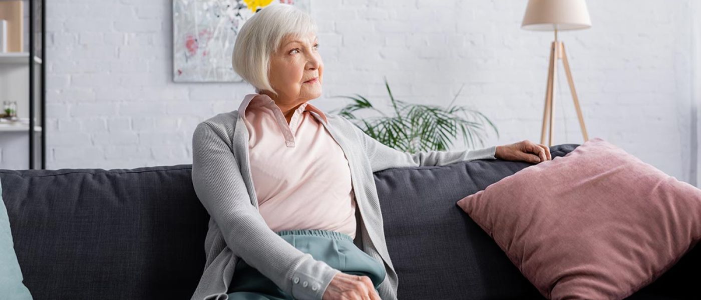 Eine Seniorin sitzt auf ihrem Sofa in ihrem Wohnzimmer, im Hintergrund hängt ein modernes Gemälde, daneben stehen eine Topfpflanze, eine Stehlampe und ein Regal | Wohnen im Alter